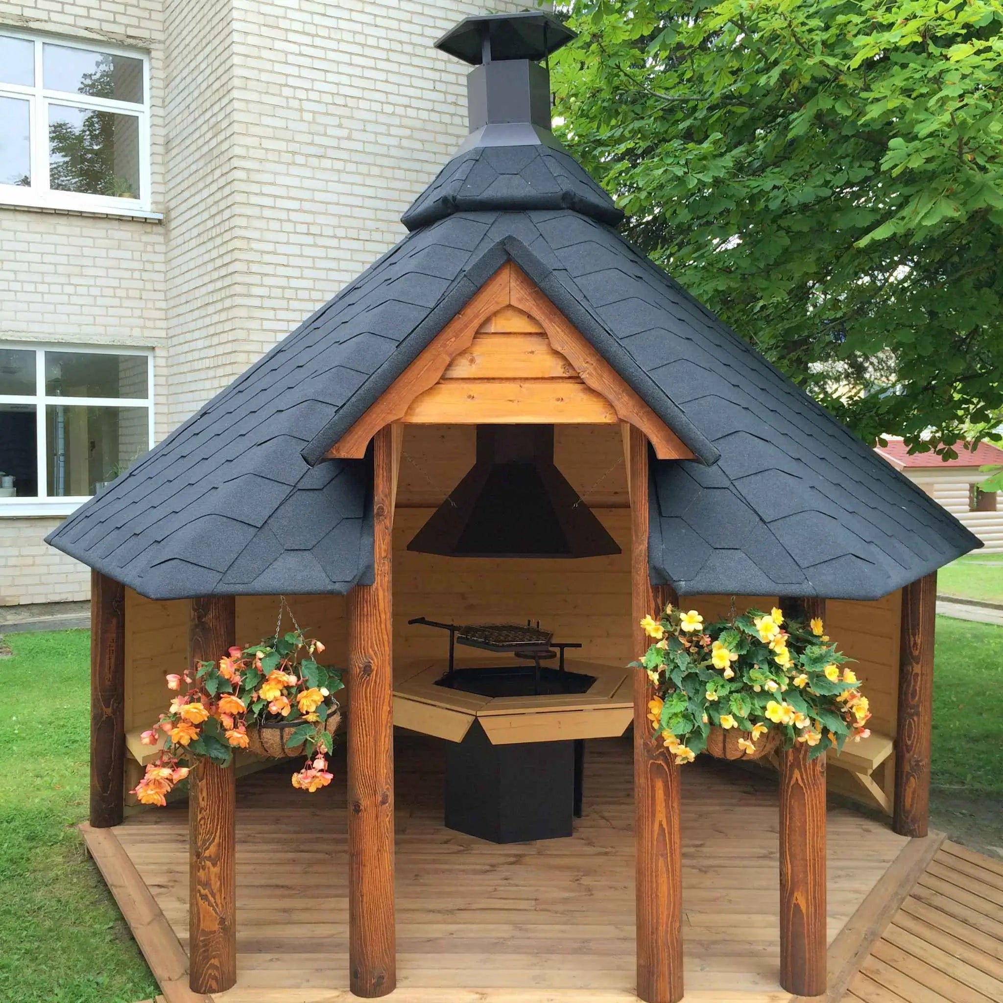 Front view of The Valhalla Shelter grill cabin, beautifully decorated with hanging flower baskets, a central barbecue grill, and traditional Nordic craftsmanship for an enchanting garden space.